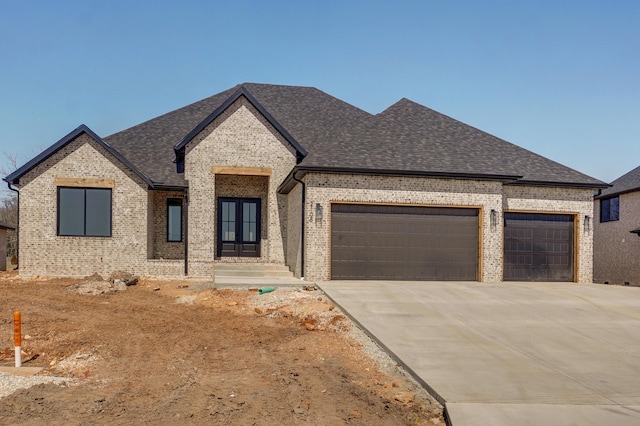 french country style house with a garage, brick siding, driveway, and roof with shingles