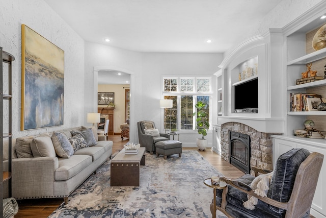 living area featuring built in shelves, arched walkways, a stone fireplace, and wood finished floors