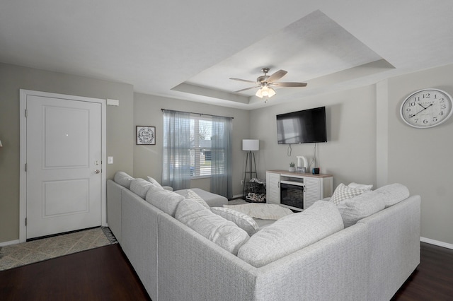 living area featuring a tray ceiling, baseboards, dark wood-type flooring, and ceiling fan