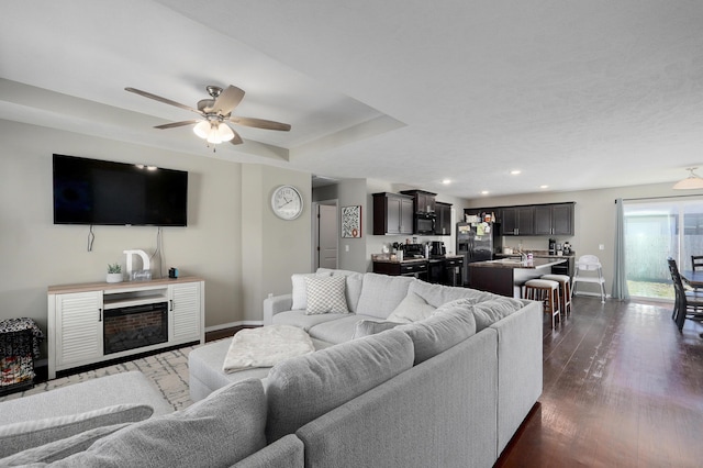 living room featuring a raised ceiling, a fireplace, baseboards, ceiling fan, and dark wood-style flooring