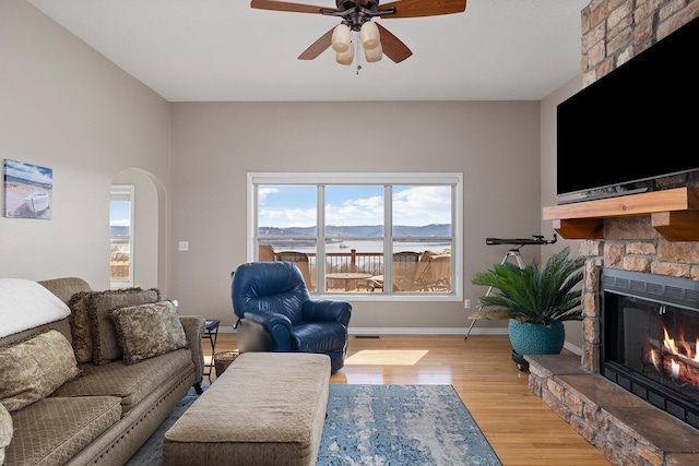 living area featuring baseboards, a fireplace, light wood-style floors, arched walkways, and a ceiling fan