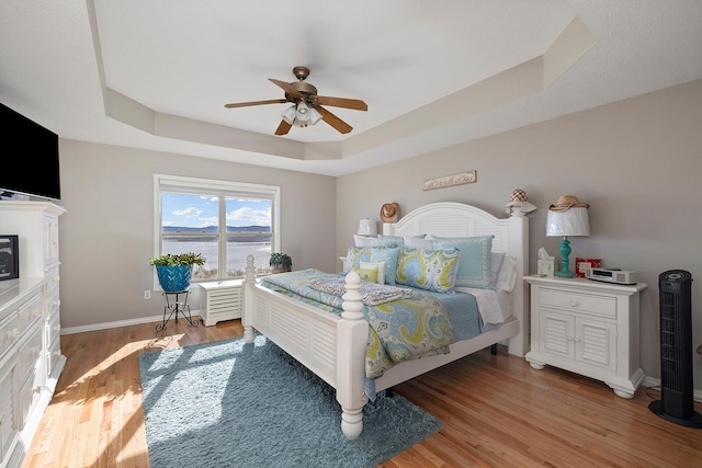 bedroom featuring baseboards, a raised ceiling, light wood-style floors, and a ceiling fan