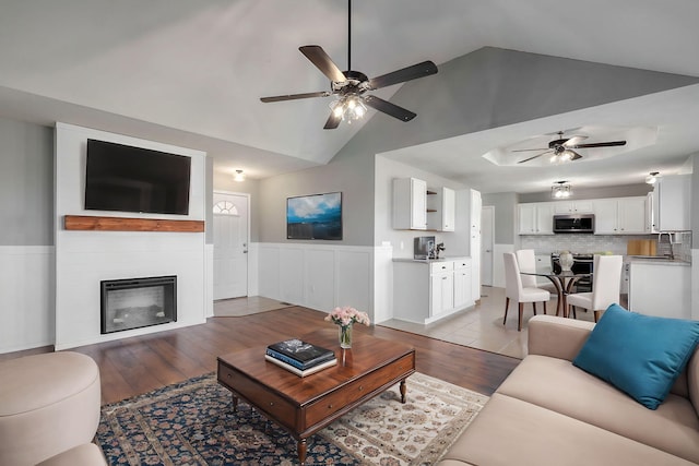 living room with light wood-type flooring, a ceiling fan, a glass covered fireplace, and wainscoting