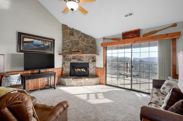 carpeted living room with visible vents, wooden walls, a stone fireplace, wainscoting, and ceiling fan