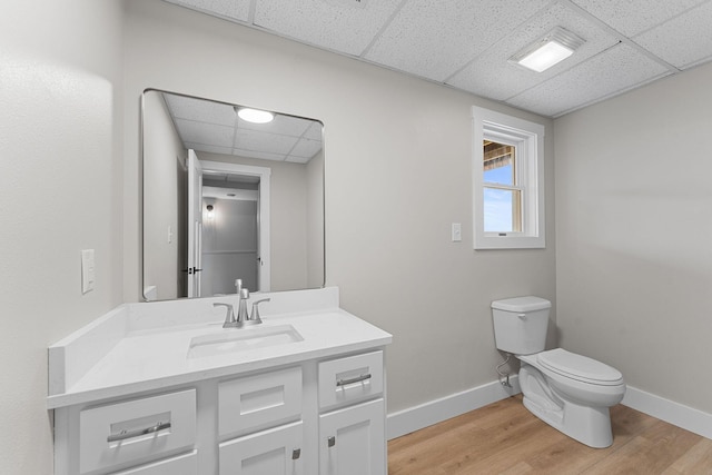 half bath with toilet, wood finished floors, baseboards, a paneled ceiling, and vanity