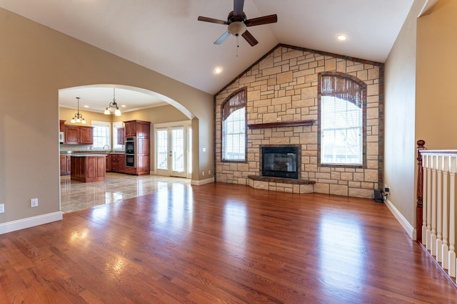 unfurnished living room featuring vaulted ceiling, a stone fireplace, light wood-style flooring, arched walkways, and a ceiling fan