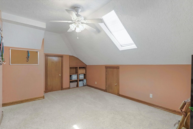 bonus room featuring a ceiling fan, a textured ceiling, vaulted ceiling with skylight, baseboards, and light colored carpet