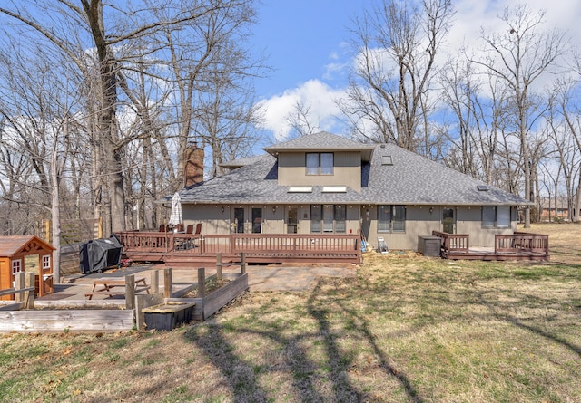 rear view of house with a wooden deck, roof with shingles, a lawn, a chimney, and an outbuilding