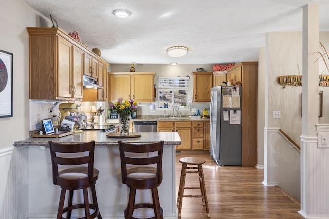 kitchen featuring under cabinet range hood, a sink, wood finished floors, stainless steel appliances, and a peninsula