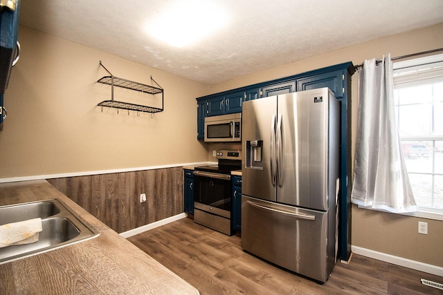 kitchen with wooden walls, a wainscoted wall, blue cabinetry, dark wood-type flooring, and appliances with stainless steel finishes