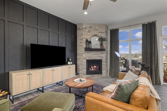 living area featuring a decorative wall, a stone fireplace, ceiling fan, and wood finished floors