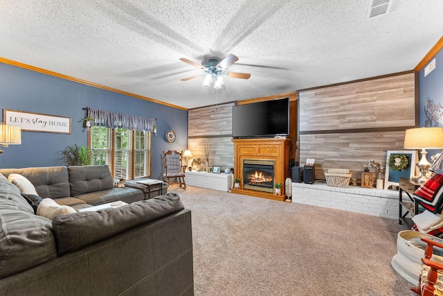 carpeted living area with visible vents, ornamental molding, a glass covered fireplace, wood walls, and ceiling fan