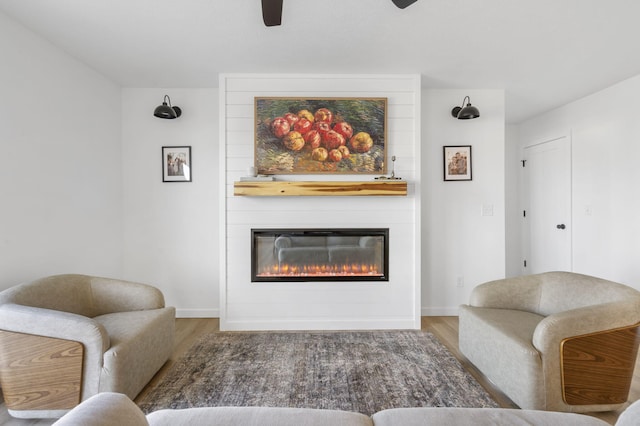 living area with a ceiling fan, wood finished floors, baseboards, and a large fireplace