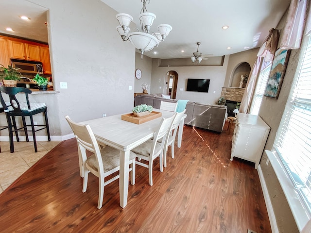 dining area with baseboards, a stone fireplace, ceiling fan with notable chandelier, wood finished floors, and arched walkways