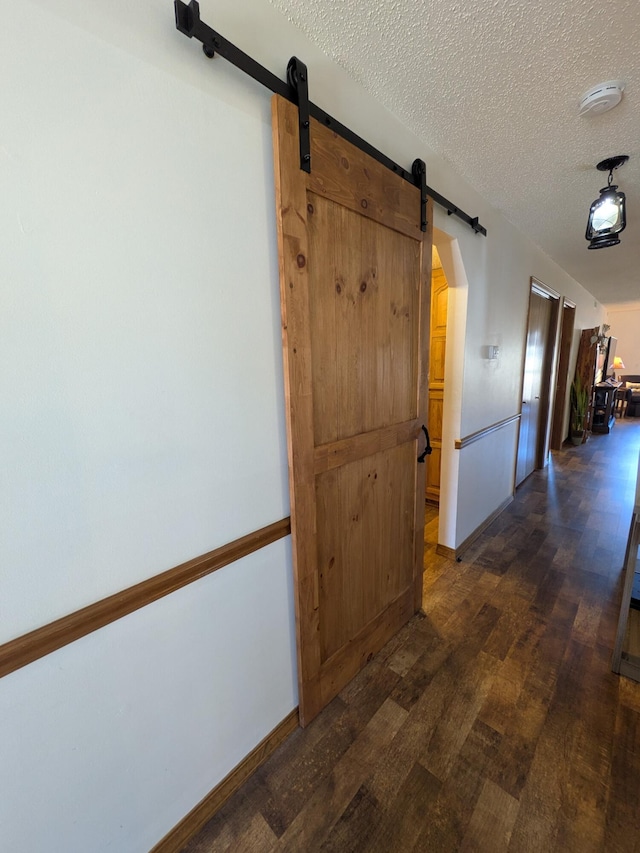 corridor featuring a textured ceiling, a barn door, arched walkways, baseboards, and dark wood-style flooring