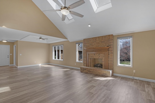 unfurnished living room featuring baseboards, a skylight, ceiling fan, a brick fireplace, and light wood-type flooring