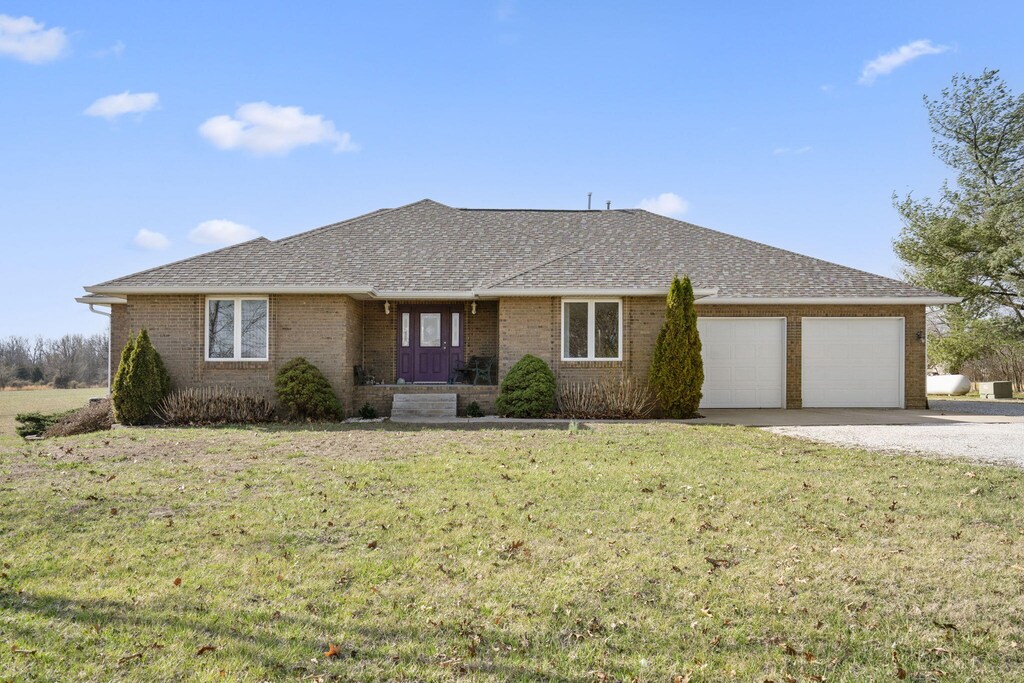 single story home featuring gravel driveway, an attached garage, brick siding, a front lawn, and a shingled roof