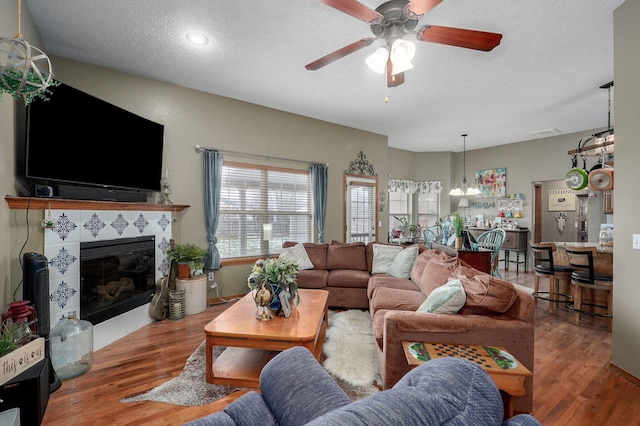 living area featuring visible vents, a tiled fireplace, ceiling fan with notable chandelier, a textured ceiling, and wood finished floors