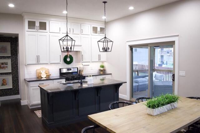 kitchen featuring dark countertops, backsplash, stainless steel range with gas stovetop, a center island with sink, and hanging light fixtures