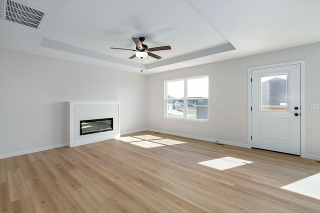 unfurnished living room featuring a ceiling fan, visible vents, light wood finished floors, baseboards, and a tray ceiling