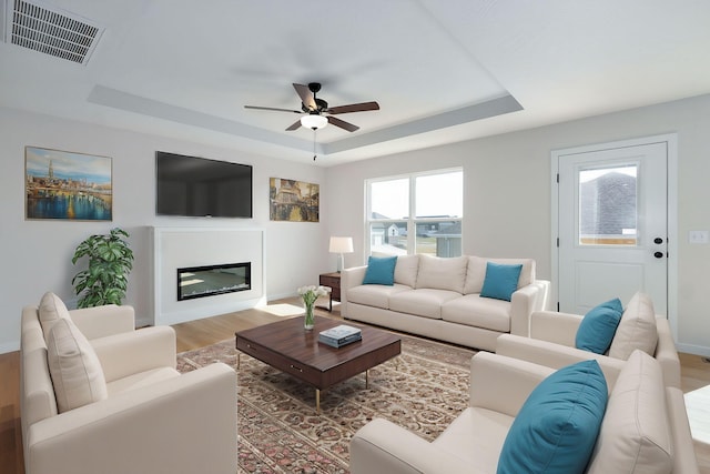 living area featuring wood finished floors, a ceiling fan, visible vents, a tray ceiling, and a glass covered fireplace