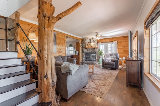 living area with stairway, a ceiling fan, wooden walls, a stone fireplace, and hardwood / wood-style floors