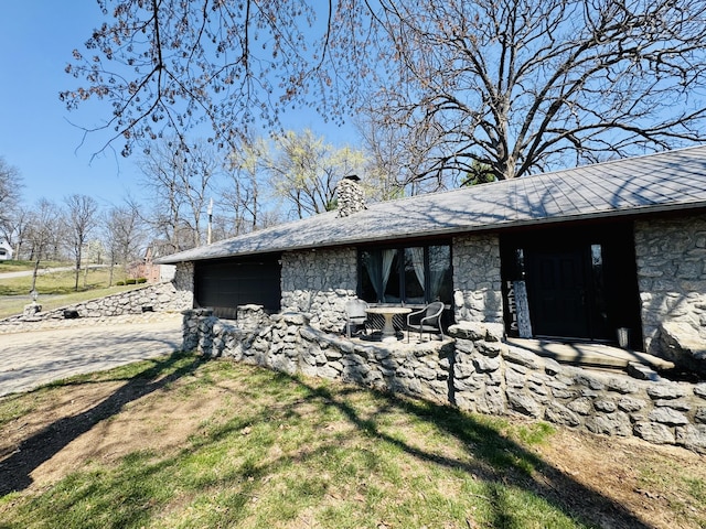 view of side of property featuring driveway, a chimney, stone siding, and an attached garage