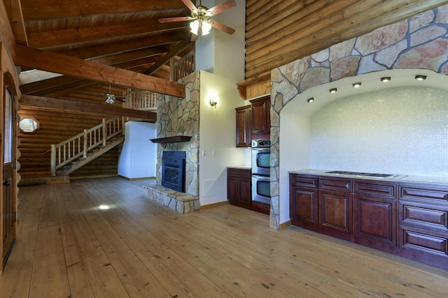 kitchen with beam ceiling, a ceiling fan, double oven, a stone fireplace, and light wood-style floors