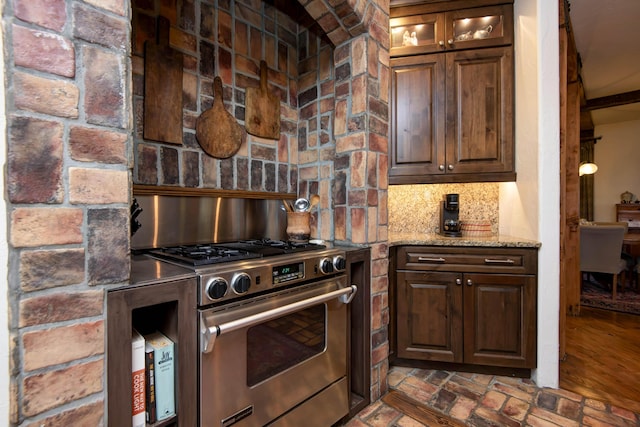 kitchen featuring decorative backsplash, stainless steel gas range, brick floor, light stone countertops, and glass insert cabinets