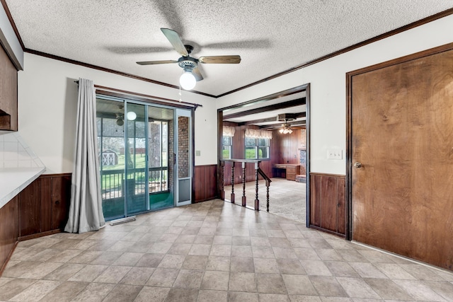 interior space with a ceiling fan, a textured ceiling, wooden walls, crown molding, and a wainscoted wall
