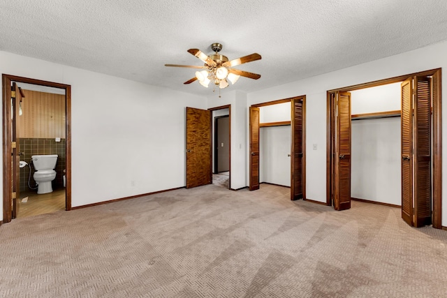 unfurnished bedroom with two closets, a textured ceiling, ensuite bath, a ceiling fan, and light carpet