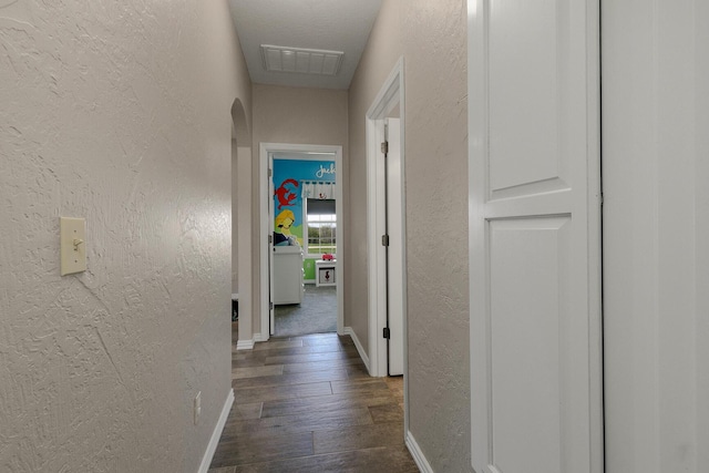 hallway with a textured wall, visible vents, dark wood-style flooring, and baseboards