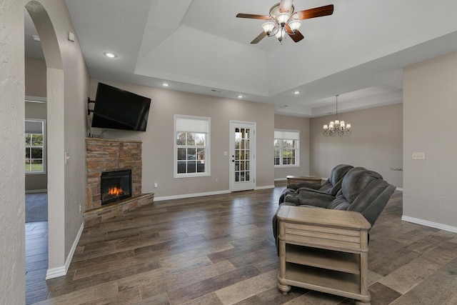 living area with a raised ceiling, wood finished floors, ceiling fan with notable chandelier, and a stone fireplace