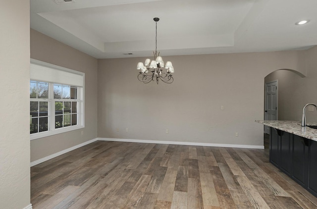 unfurnished dining area with baseboards, arched walkways, a chandelier, a tray ceiling, and dark wood-type flooring