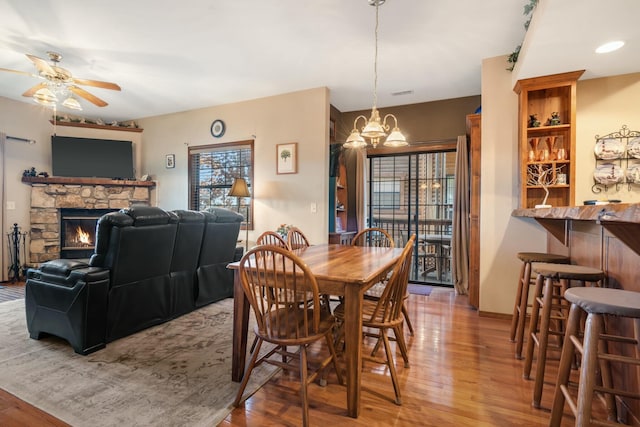 dining area with ceiling fan with notable chandelier, a dry bar, visible vents, a stone fireplace, and wood finished floors