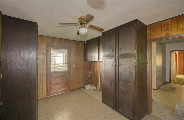 interior space featuring baseboards, wooden walls, a textured ceiling, a ceiling fan, and dark brown cabinets