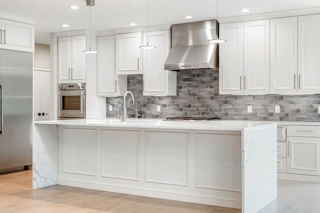 kitchen with appliances with stainless steel finishes, wall chimney range hood, an island with sink, white cabinetry, and backsplash