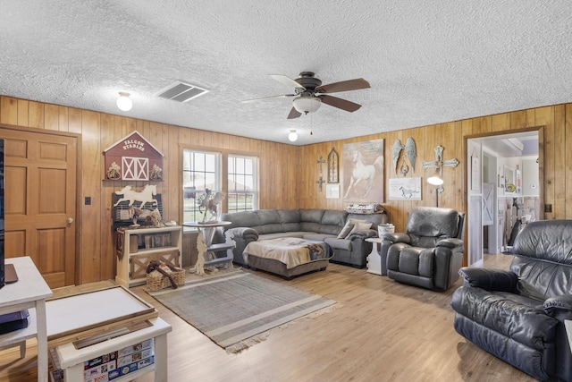 living area with a textured ceiling, light wood-type flooring, visible vents, a ceiling fan, and wood walls