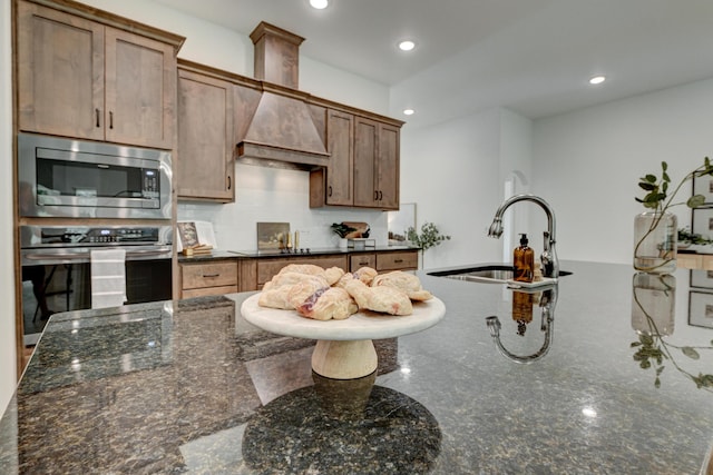 kitchen featuring dark stone counters, a sink, custom range hood, backsplash, and appliances with stainless steel finishes