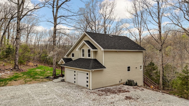 view of side of property featuring stairway, gravel driveway, central AC, roof with shingles, and a garage