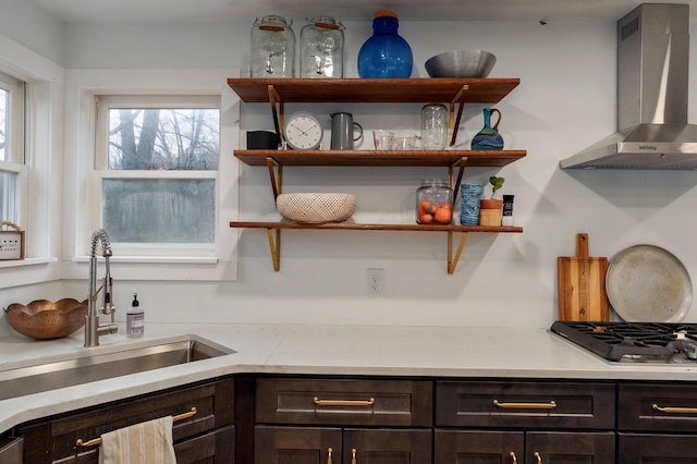 kitchen with stainless steel gas cooktop, a sink, dark brown cabinetry, wall chimney range hood, and light countertops