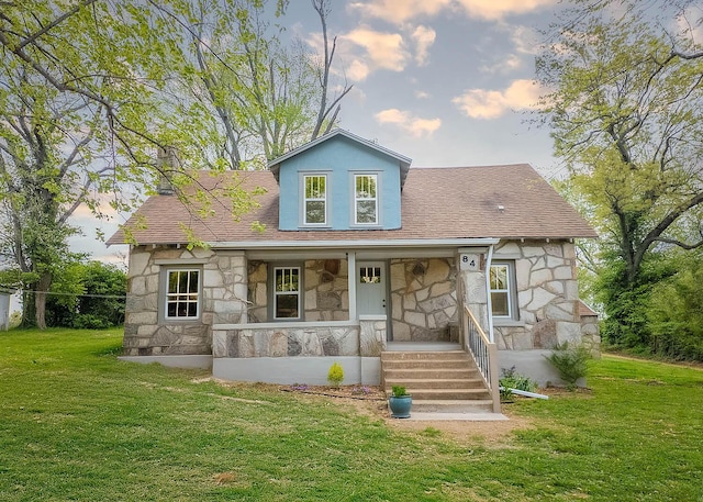 back of property at dusk with covered porch, a yard, a shingled roof, and stone siding