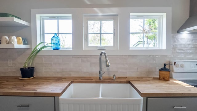 kitchen featuring wall chimney range hood, open shelves, decorative backsplash, a sink, and butcher block counters