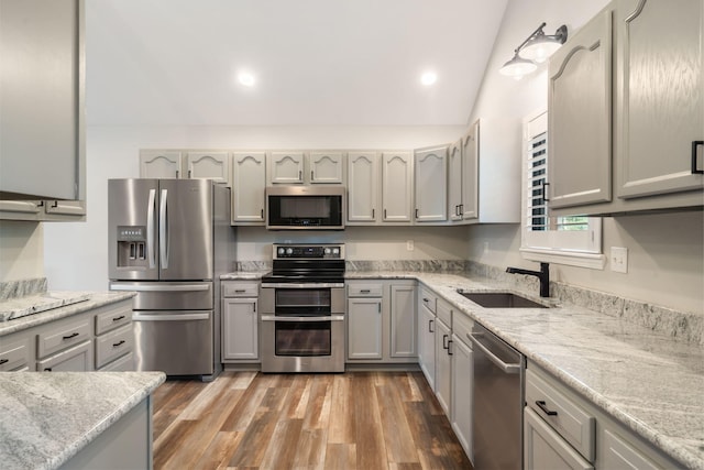 kitchen featuring appliances with stainless steel finishes, vaulted ceiling, wood finished floors, a sink, and light stone counters