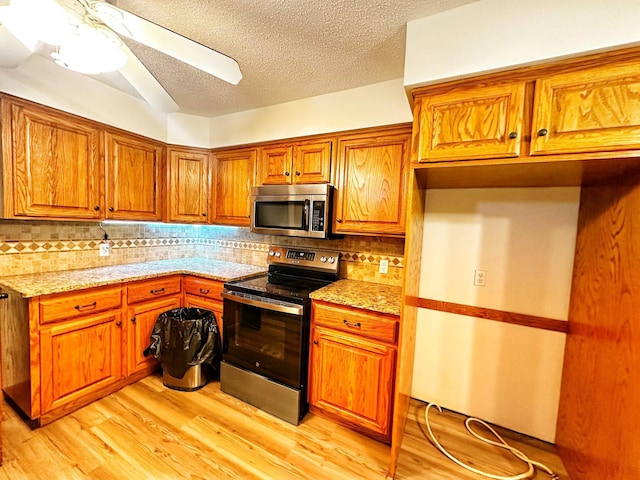 kitchen with appliances with stainless steel finishes, light wood-type flooring, light stone countertops, brown cabinetry, and backsplash