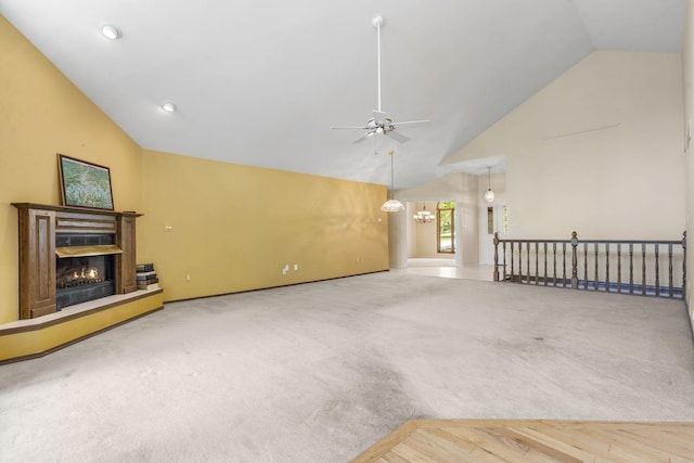 unfurnished living room featuring carpet flooring, high vaulted ceiling, a warm lit fireplace, and ceiling fan with notable chandelier