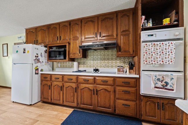 kitchen featuring white appliances, under cabinet range hood, brown cabinetry, and light countertops