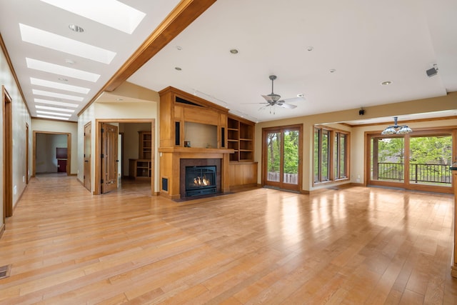 unfurnished living room with built in features, a fireplace, ceiling fan with notable chandelier, lofted ceiling with skylight, and light wood-style floors