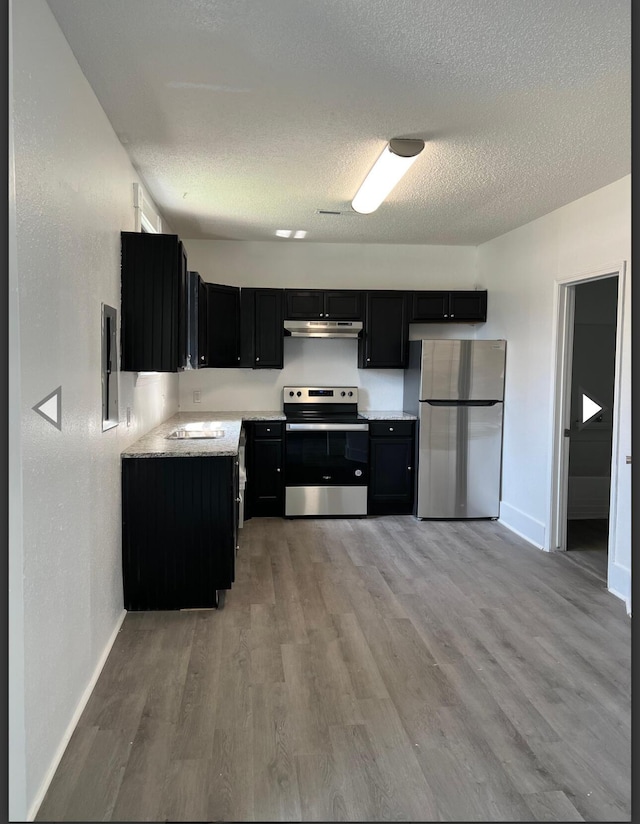 kitchen with appliances with stainless steel finishes, light wood finished floors, dark cabinetry, a textured ceiling, and under cabinet range hood