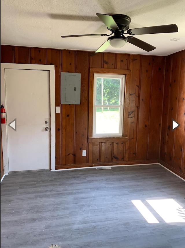 unfurnished room with a textured ceiling, wood walls, a ceiling fan, wood finished floors, and electric panel